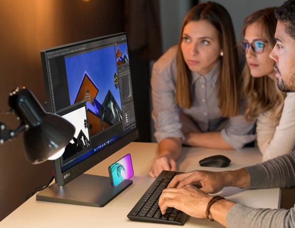 Three business analysts huddled around a ThinkCentre Neo 50a all-in-one PC on a desk, with phone (sold separately) in phone holder, plus wireless mouse & keyboard (both sold separately)